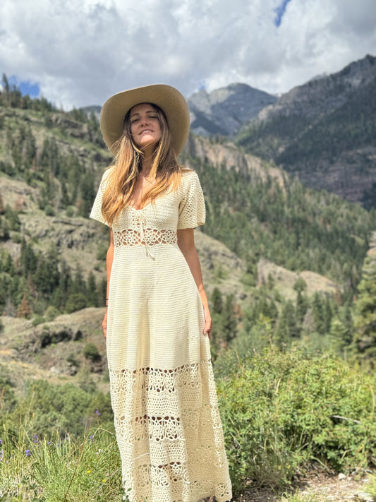 A woman wearing a cream-colored crochet maxi dress and a wide-brimmed straw hat stands outdoors in a mountain landscape with green trees and rocky peaks under a cloudy sky.